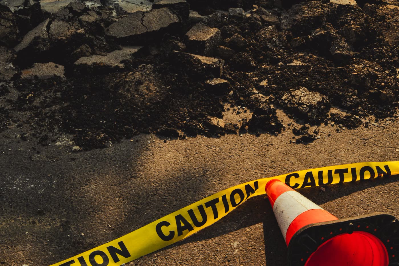 A broken pavement with rubble, caution tape and a traffic cone